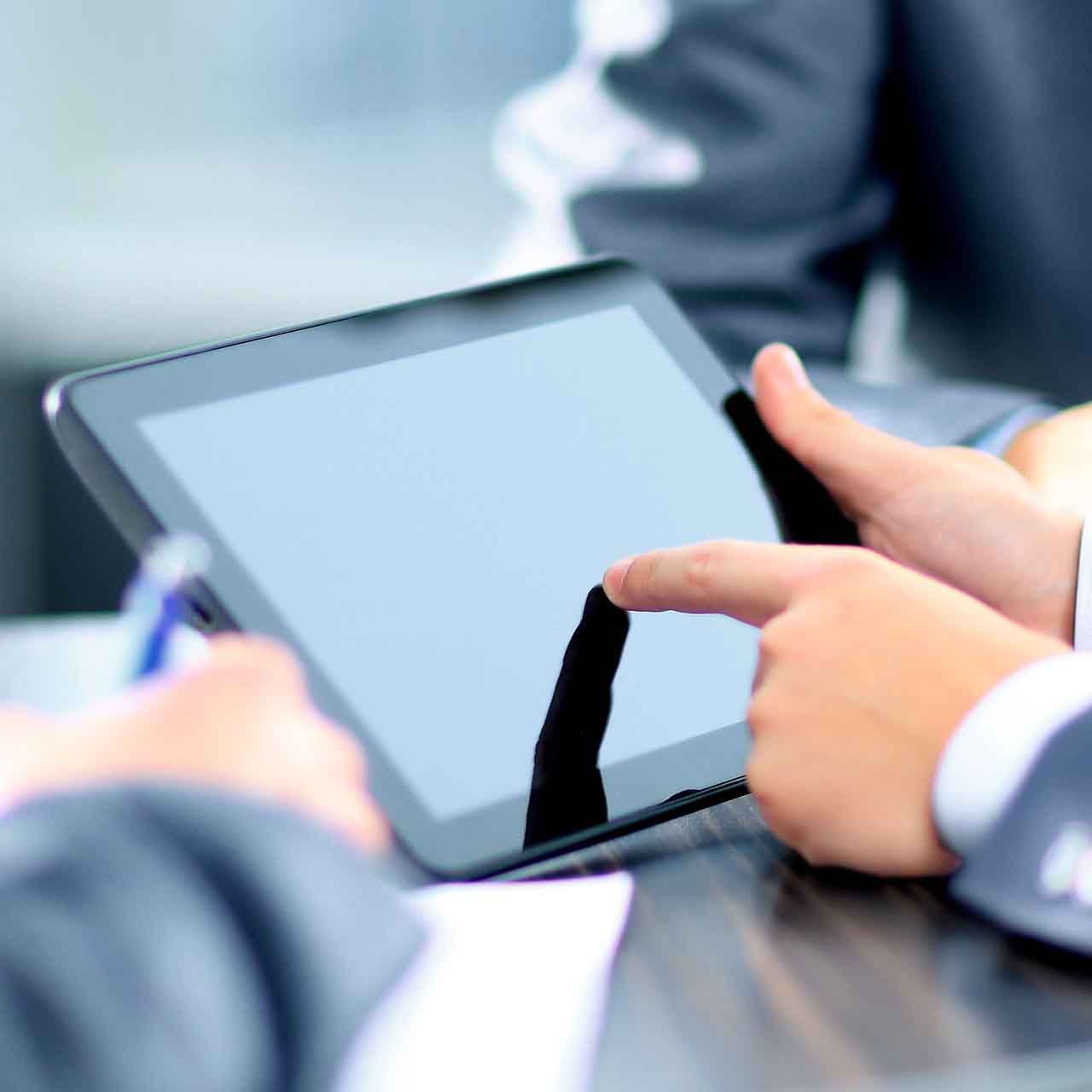 Close-up of business professionals in formal attire using a digital tablet during a meeting, with one person pointing at the screen and another holding a pen.